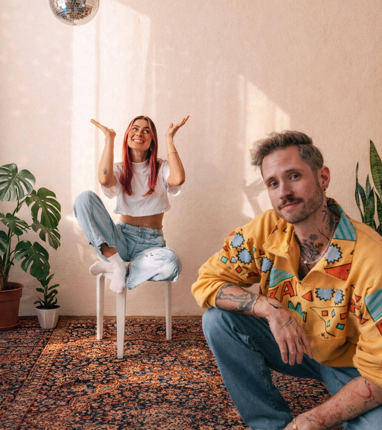 kitsch and SASH, a DJ duo, sitting in a cozy studio with plants and a disco ball, smiling and relaxed