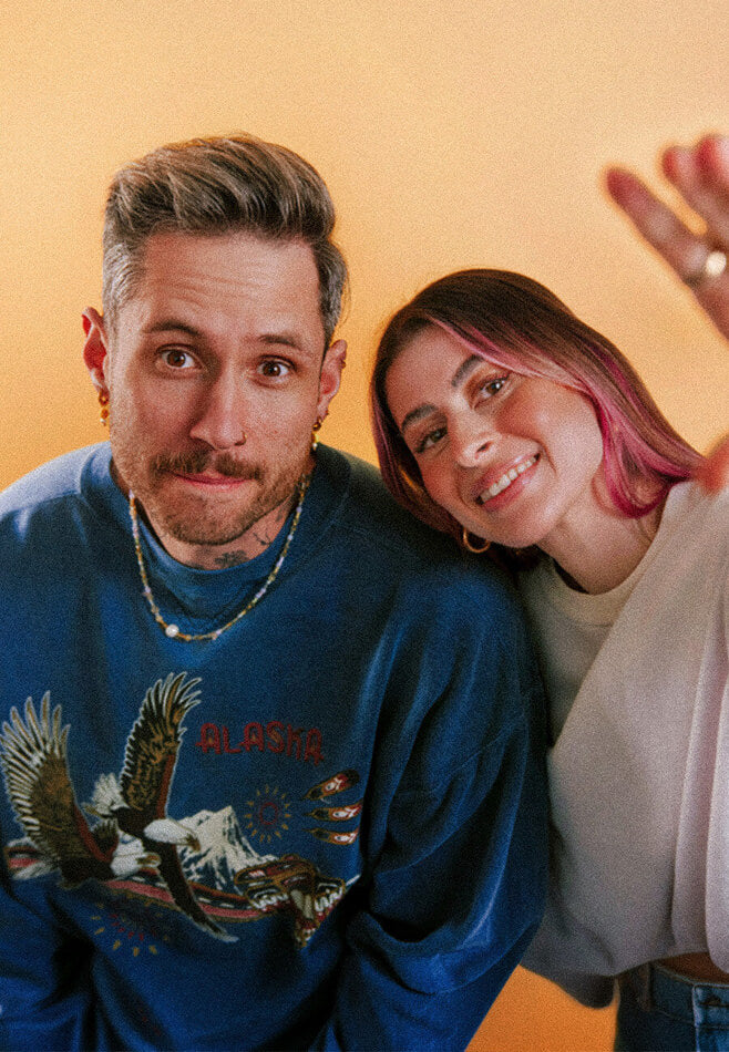 DJ Duo kitsch and SASH posing closely together and smiling in a friendly studio setting in front of a yellow background
